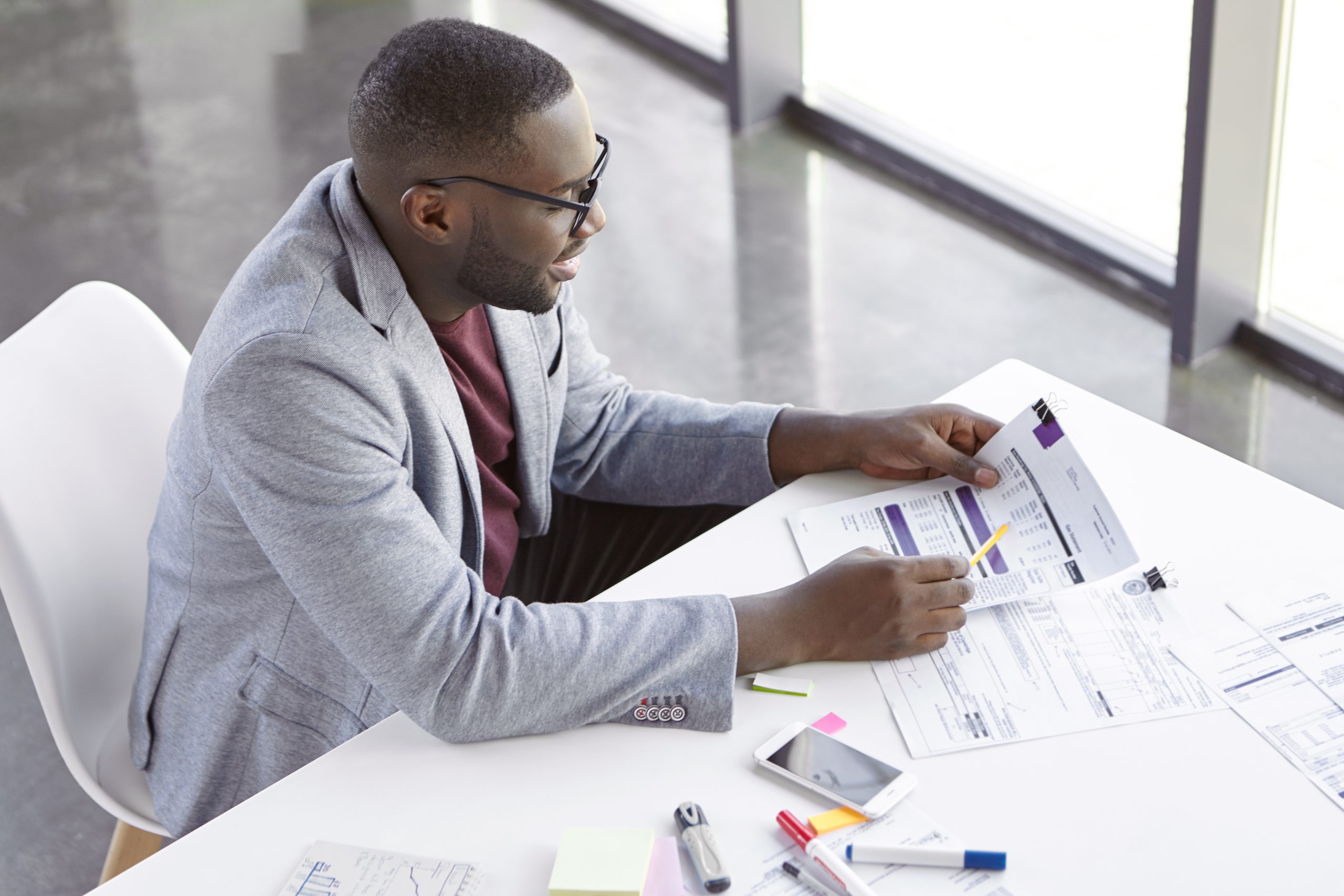 Top view of dark skinned African male copywriter or enterpreneur looks attentively at documents, studies net profit of his company, sits at cozy cabinet, in private office, does paper work alone